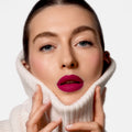 A person wearing a white turtleneck sweater pulled up to their chin, featuring Sisley Paris Phyto-Rouge Velvet lipstick in a bold, matte magenta hue.