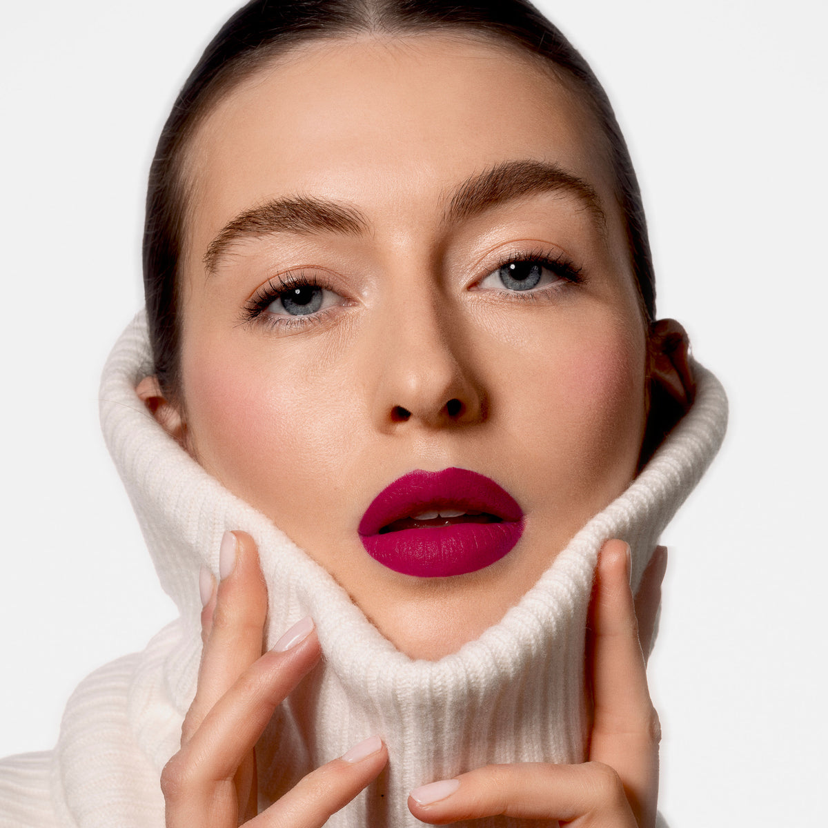 A person wearing a white turtleneck sweater pulled up to their chin, featuring Sisley Paris Phyto-Rouge Velvet lipstick in a bold, matte magenta hue.