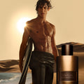A shirtless surfer with wet hair stands on a desert beach at sunset holding a surfboard next to a bottle of Tom Ford Oud Minerale Eau de Parfum, capturing a sense of adventure.