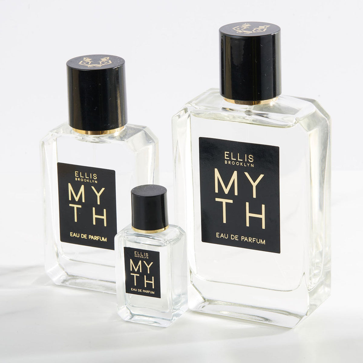 Three rectangular glass bottles of MYTH Eau de Parfum in varying sizes, each with a black cap and a black label with gold lettering, carrying notes of jasmine petals and white cedarwood.