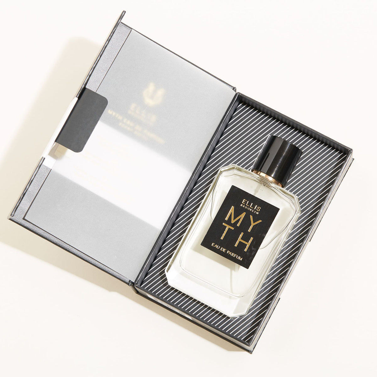 A rectangular bottle of MYTH Eau de Parfum with a black cap and a black label with gold text rests inside an open gift box containing notes of liquid musks and jasmine petals.