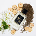 A rectangular MYTH Eau de Parfum glass bottle with a black label surrounded by jasmine petals, white cedarwood slices, and scattered seeds on a white surface.