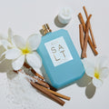 A light blue rectangular bottle of Ellis Brooklyn Salt Eau de Parfum, evoking salty skin, surrounded by white tropical florals, wooden sticks, and coarse sea salt.