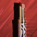 Phyto-Rouge Shine lipstick with a dark red tip and a gold case featuring a lavender zebra-stripe pattern, set against a red-tinted background.