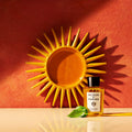 A bottle of Acqua di Parma Buongiorno Eau de Parfum with a sprig of basil in front of it, placed before a decorative yellow sun-shaped wall plaque.