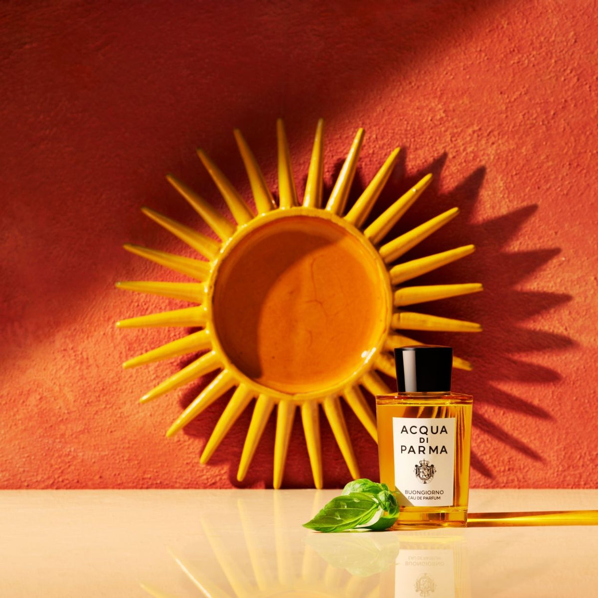 A bottle of Acqua di Parma Buongiorno Eau de Parfum with a sprig of basil in front of it, placed before a decorative yellow sun-shaped wall plaque.