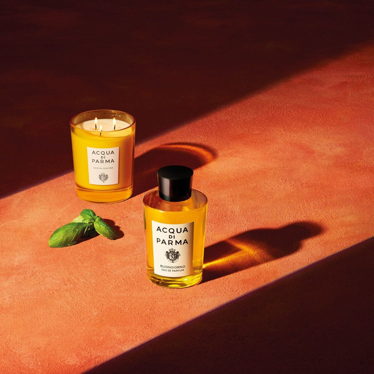 An Acqua di Parma Buongiorno Eau de Parfum bottle and a scented candle in a yellow glass jar, both labeled, sit next to a sprig of fresh basil on an orange surface to evoke spring in Tuscany.