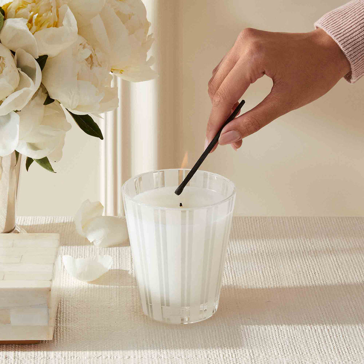 A hand uses a long match to light the wick of a striped glass Nest New York Grapefruit Candle set next to a bouquet of white flowers.