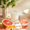 A Nest New York Grapefruit candle in a fluted glass jar, next to a halved grapefruit, a small pitcher of white flowers, and a glass of water on a tray.