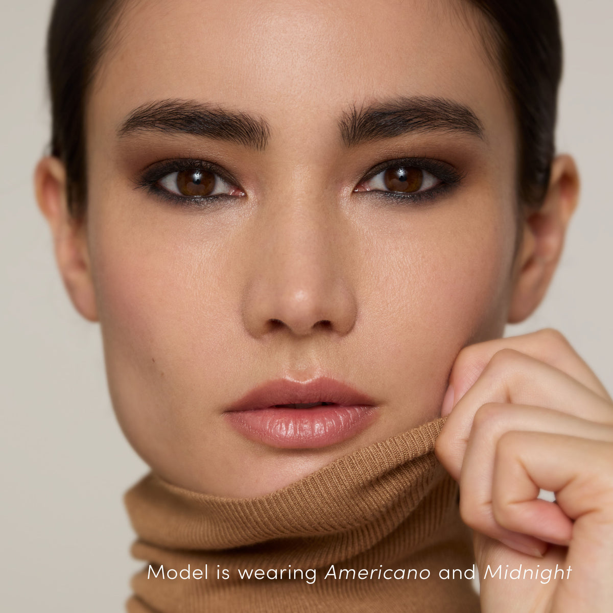 Close-up of a person wearing ColorLuxe Eye Shadow Stick in dark brown and black, with text at the bottom reading Model is wearing Americano and Midnight.