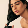A person with dark, medium to thick hair looks down while applying Christophe Robin Night Recovery Monoï Oil with White Lotus Flower to a section of their hair.