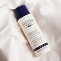 A white bottle of Christophe Robin Night Recovery Monoï Oil with White Lotus Flower featuring a dark blue cap, resting on crumpled white fabric.