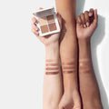 A hand holds a Lune+Aster Eyeshadow Quad above three arms displaying swatches of the four shades on different skin tones.