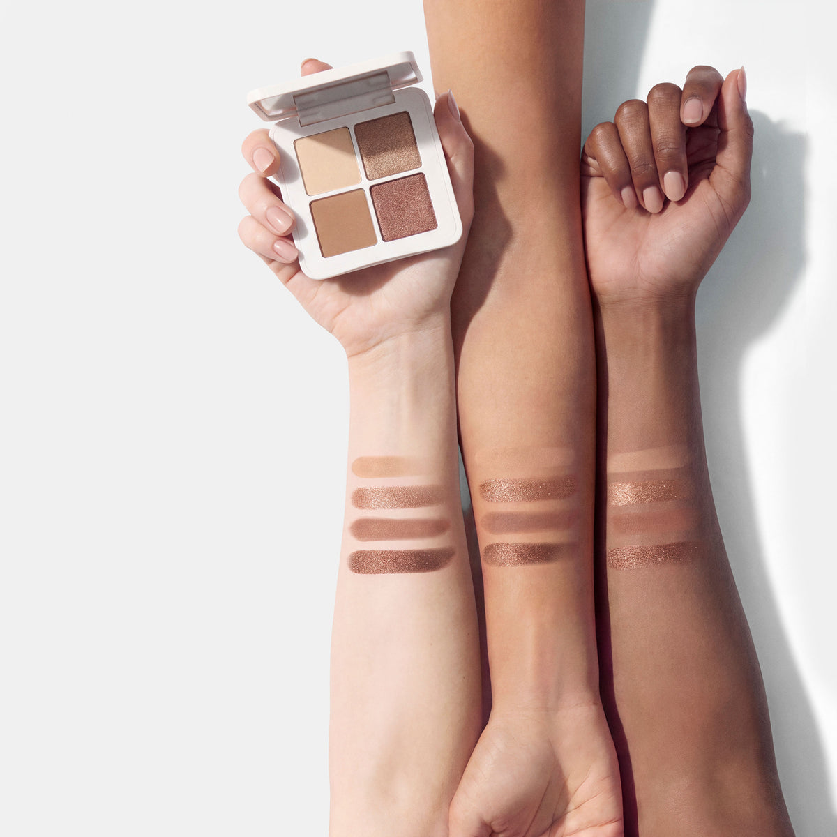 A hand holds a Lune+Aster Eyeshadow Quad above three arms displaying swatches of the four shades on different skin tones.