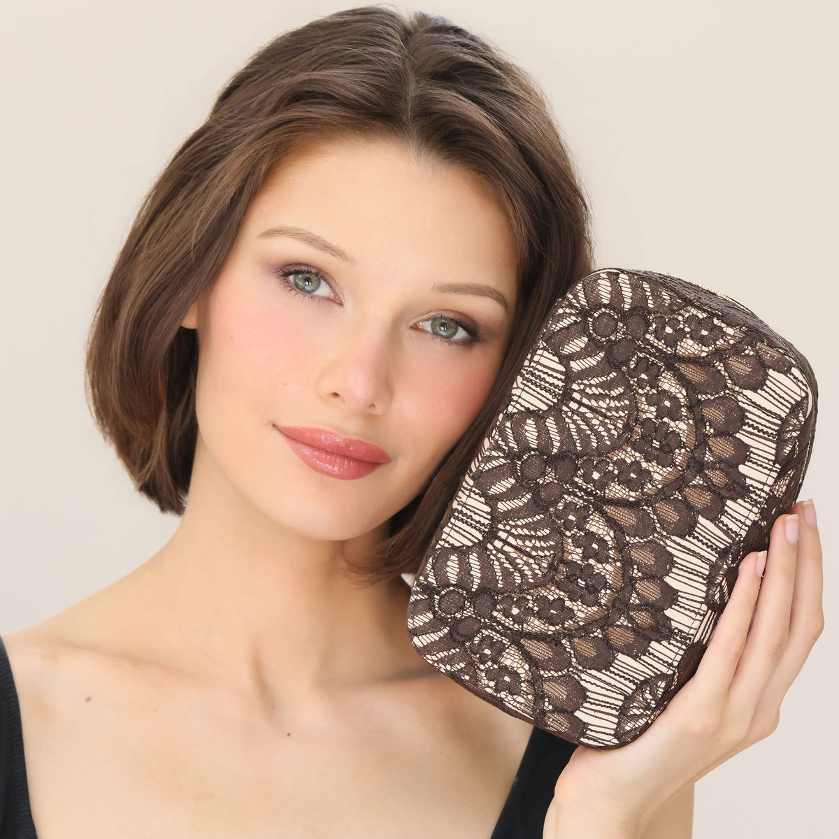 A woman holds a small, rectangular The Bluemercury Exclusive Holiday Makeup Wardrobe (Limited Edition) with a dark brown floral lace overlay, capturing a festive holiday mood.