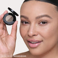 A person holds an open Anastasia Beverly Hills Brow Powder Duo compact next to their face to achieve a natural-looking finish.